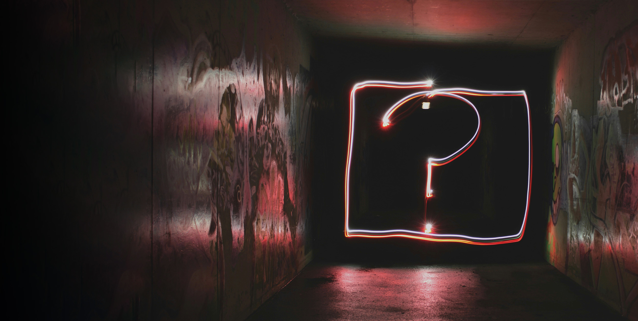 Ett frågetecken ritat med ljus i en mörk gångtunnel med graffiti på väggarna.